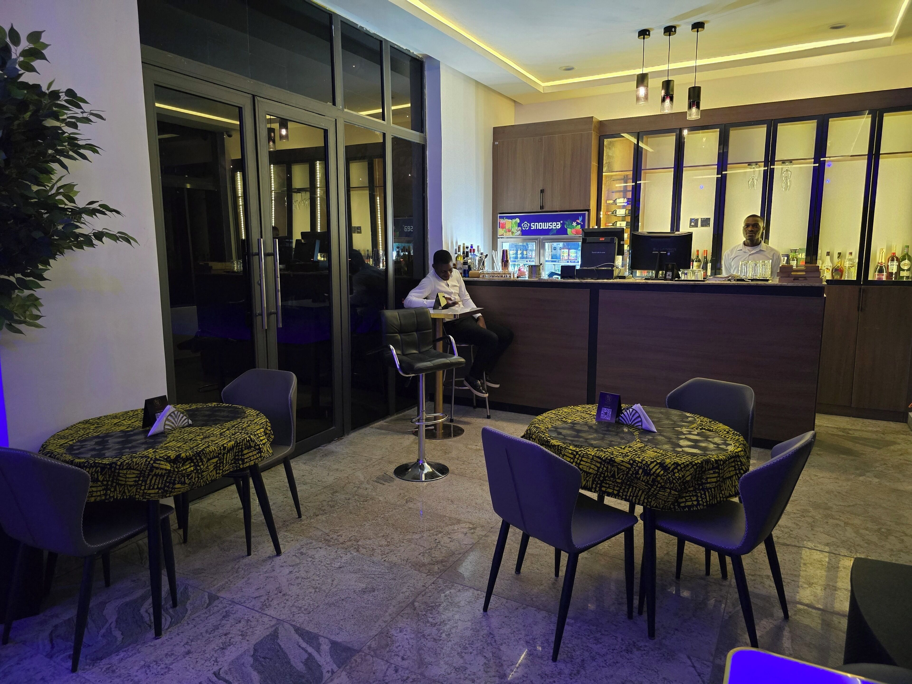 Een moderne bar met een toonbank, barkrukken en een zithoek met ronde tafels en stoelen.