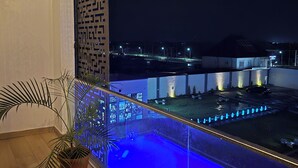 A balcony with a pool, potted plants, and a view of a lit-up area with fountains and a car. - AIKU LUXURY HOTEL & RESORTS  (Uyo)