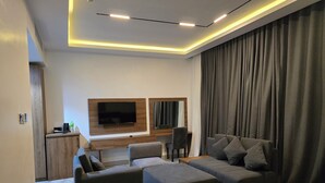 A modern hotel room with a large bed, a sofa, a desk with a flat-screen TV, and a large window with curtains. - AIKU LUXURY HOTEL & RESORTS  (Uyo)