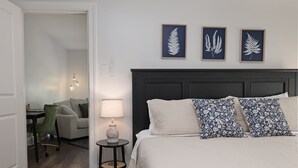 Quarto bem arrumado com uma cama grande, mesa de cabeceira com abajur, cadeira e escrivaninha com sua respectiva cadeira.