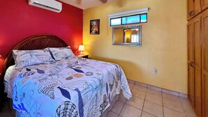 A bedroom with a bed, a ceiling fan, an air conditioner, a bedside lamp, a mirror, and wooden cabinets.