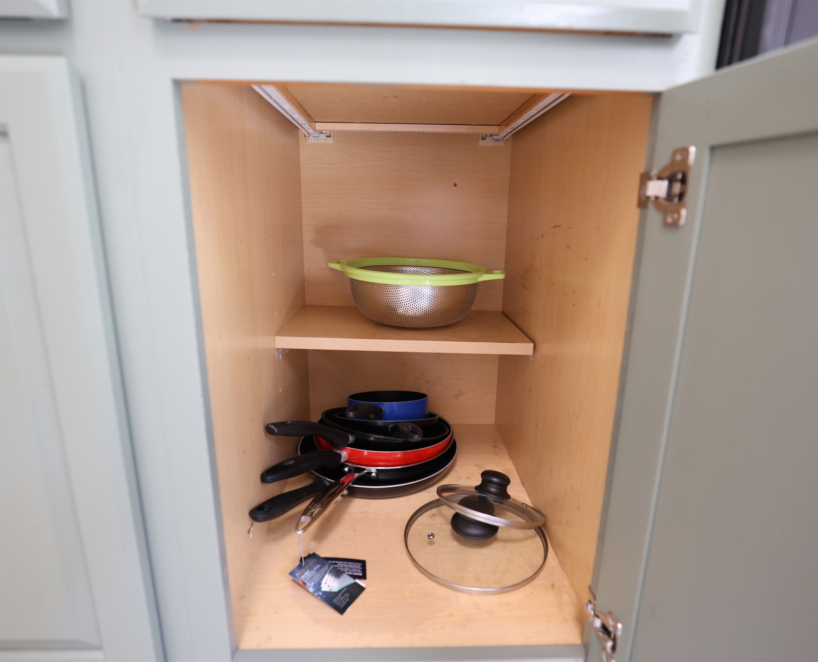 A kitchen cabinet with a wooden interior, containing a colander, a frying pan, a pot, a spatula, and a lid.