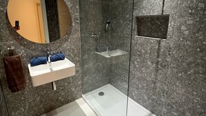 Uma casa de banho moderna com cabine de duche em vidro, lavatório branco com espelho e parede revestida a mármore.