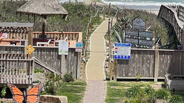 Uma trilha que leva a uma área de praia, com cercas de madeira, placas indicando acesso à praia e uma estrutura com telhado de palha.