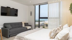 Un dormitorio moderno con una cama grande, un sofá gris, un televisor montado en la pared y un ventanal con vistas a la playa.