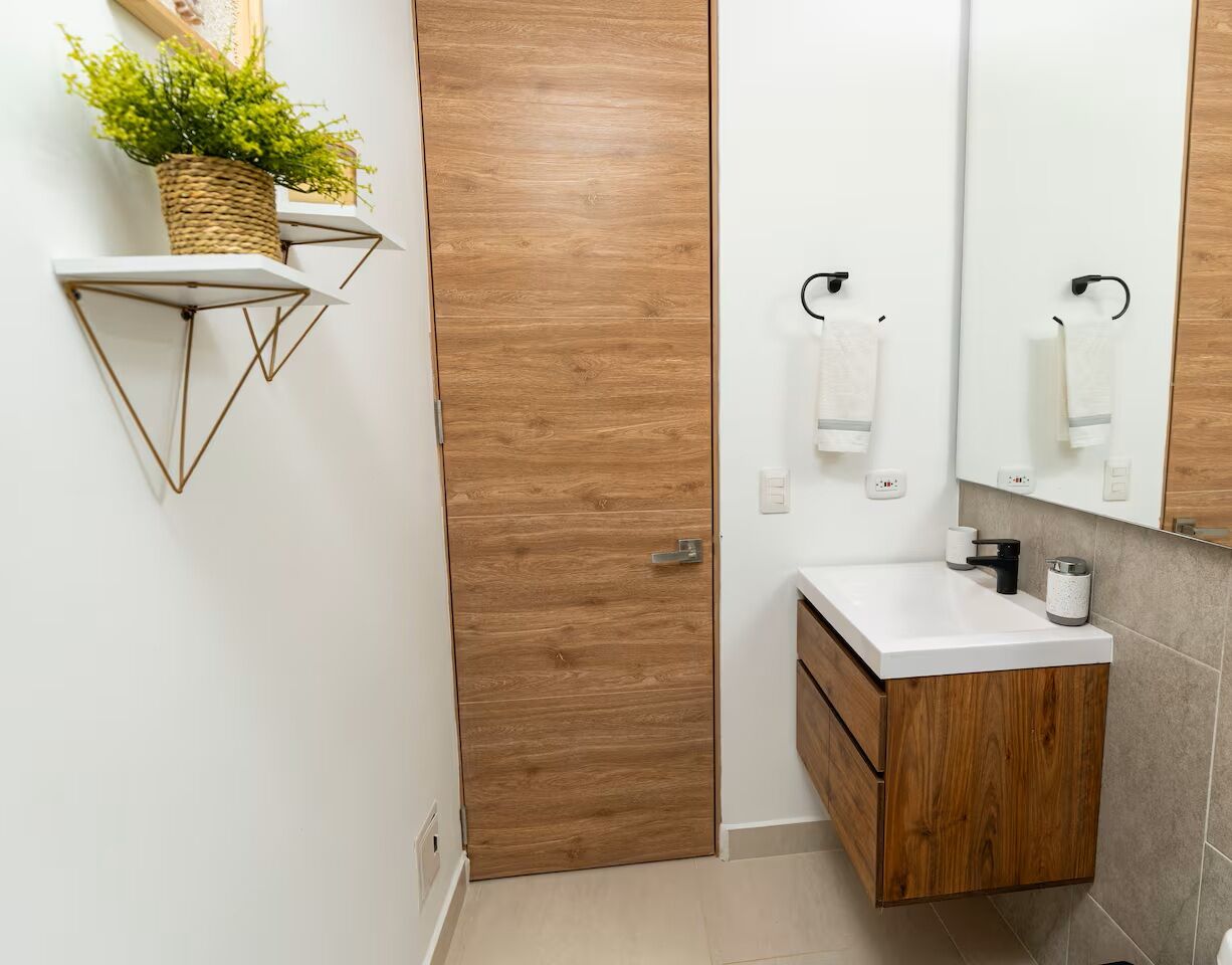 Une salle de bain avec une porte en bois, un lavabo blanc avec un meuble en bois, un miroir et une étagère murale agrémentée d’une plante.
