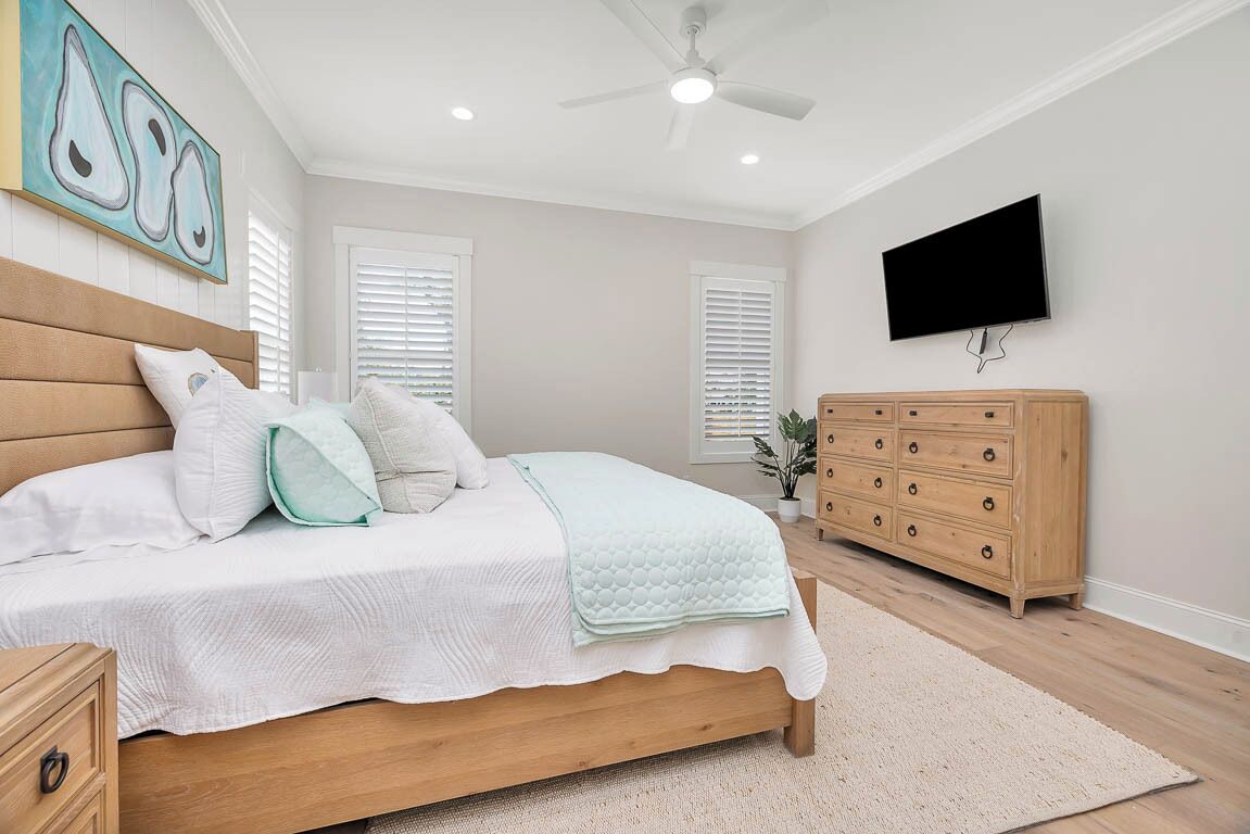 Une chambre avec un lit en bois, du linge de lit blanc, une commode en bois, un ventilateur de plafond et un téléviseur fixé au mur.