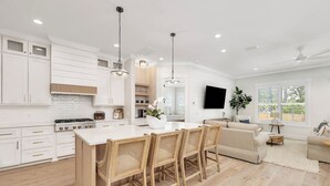 A modern kitchen with white cabinetry, a central island with chairs, a dining table, and a TV mounted on the wall.