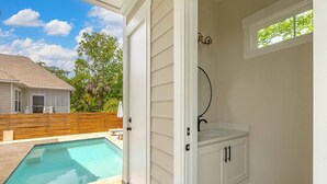 A bathroom with a white vanity, a shower, and a window with a view of a backyard swimming pool.