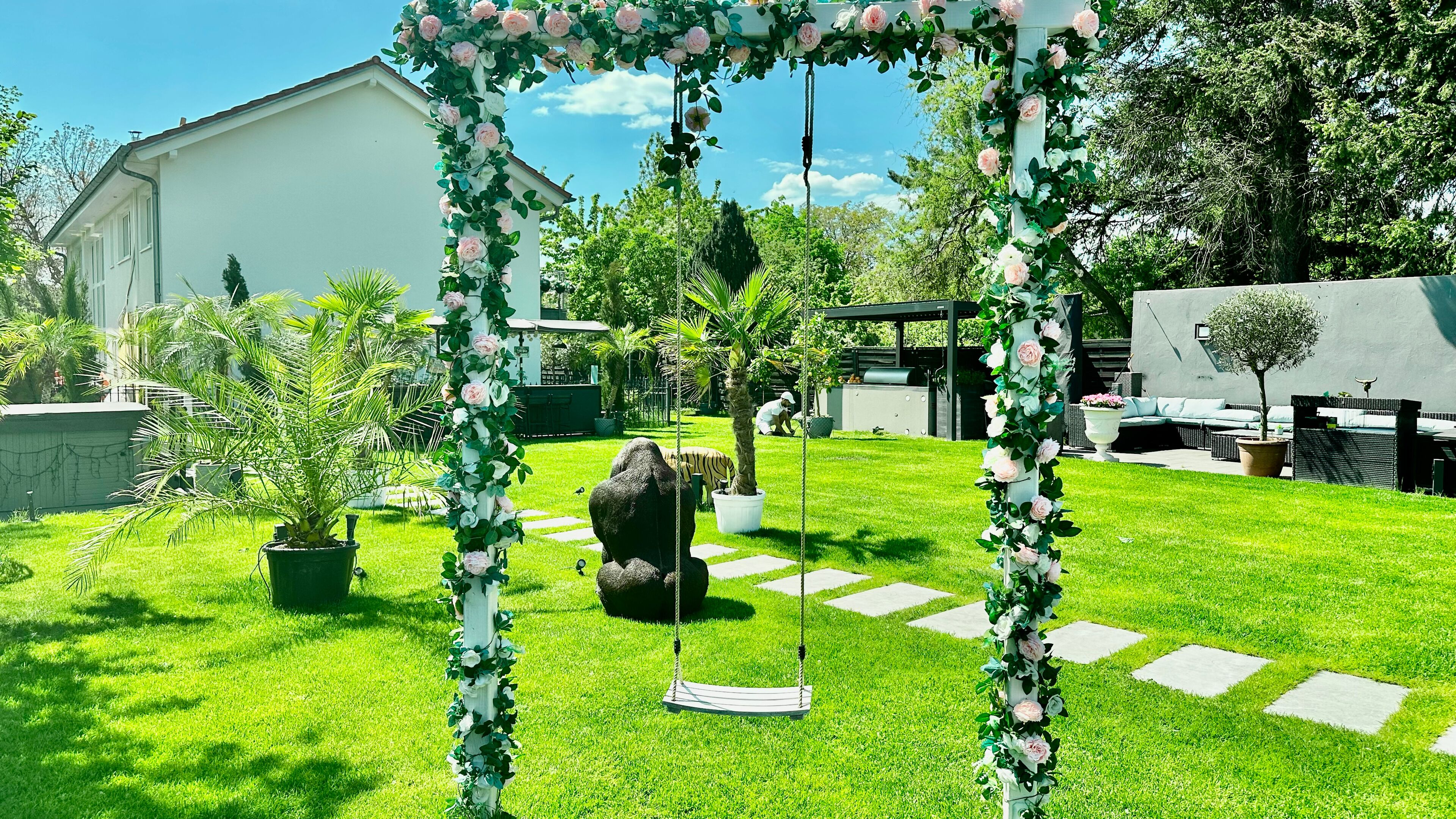 A garden with a swing set, a large rock, and a white building in the background.