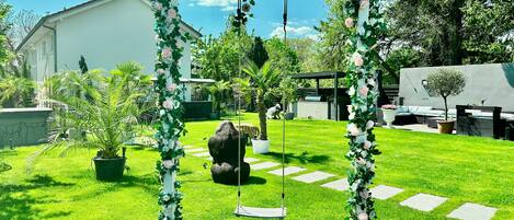 A garden with a swing set, a large rock, and a white building in the background.