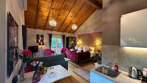 A modern hotel room with a wooden ceiling, a bed with a red bedspread, a sitting area with a sofa and armchairs, a dining table with a vase of flowers, and a kitchen area with a sink and kettle. - Villa Tropicana (Teltow)