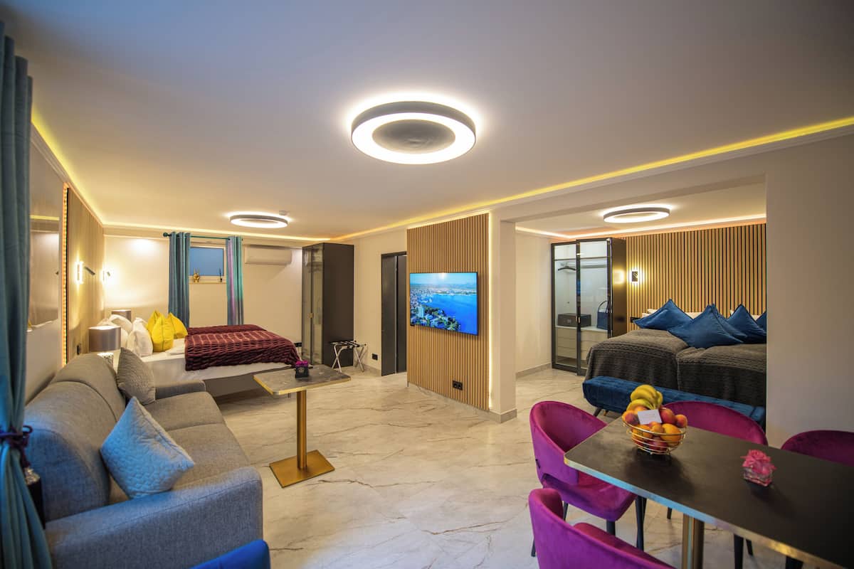 A modern hotel room with a sofa, a dining table with chairs, a TV, and a balcony with a view.