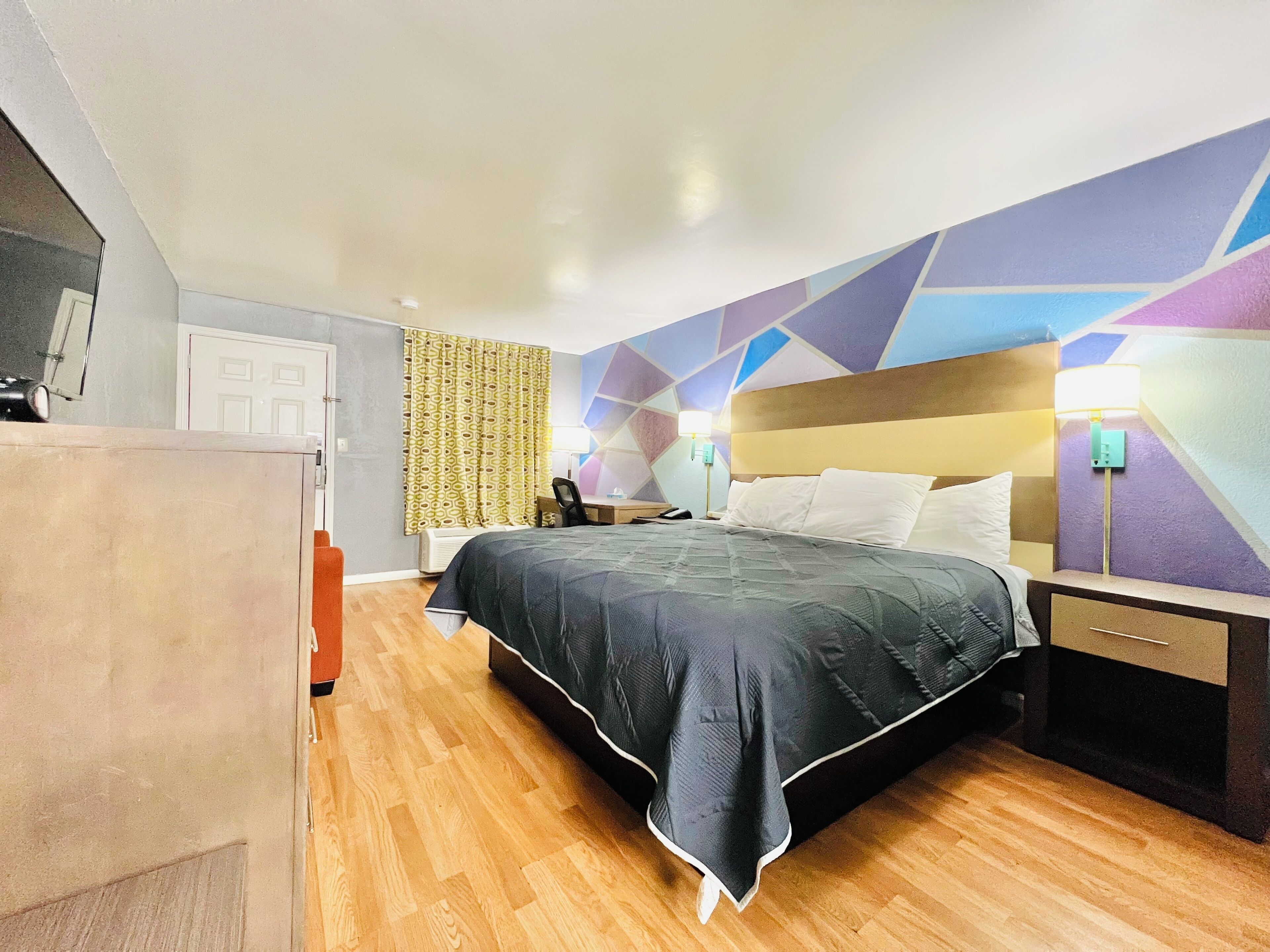 A hotel room with a bed, bedside table, lamp, and a patterned curtain.