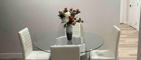 Un comedor con una mesa de vidrio rodeada por cuatro sillas blancas, un jarrón con flores y un cuadro abstracto moderno en la pared.