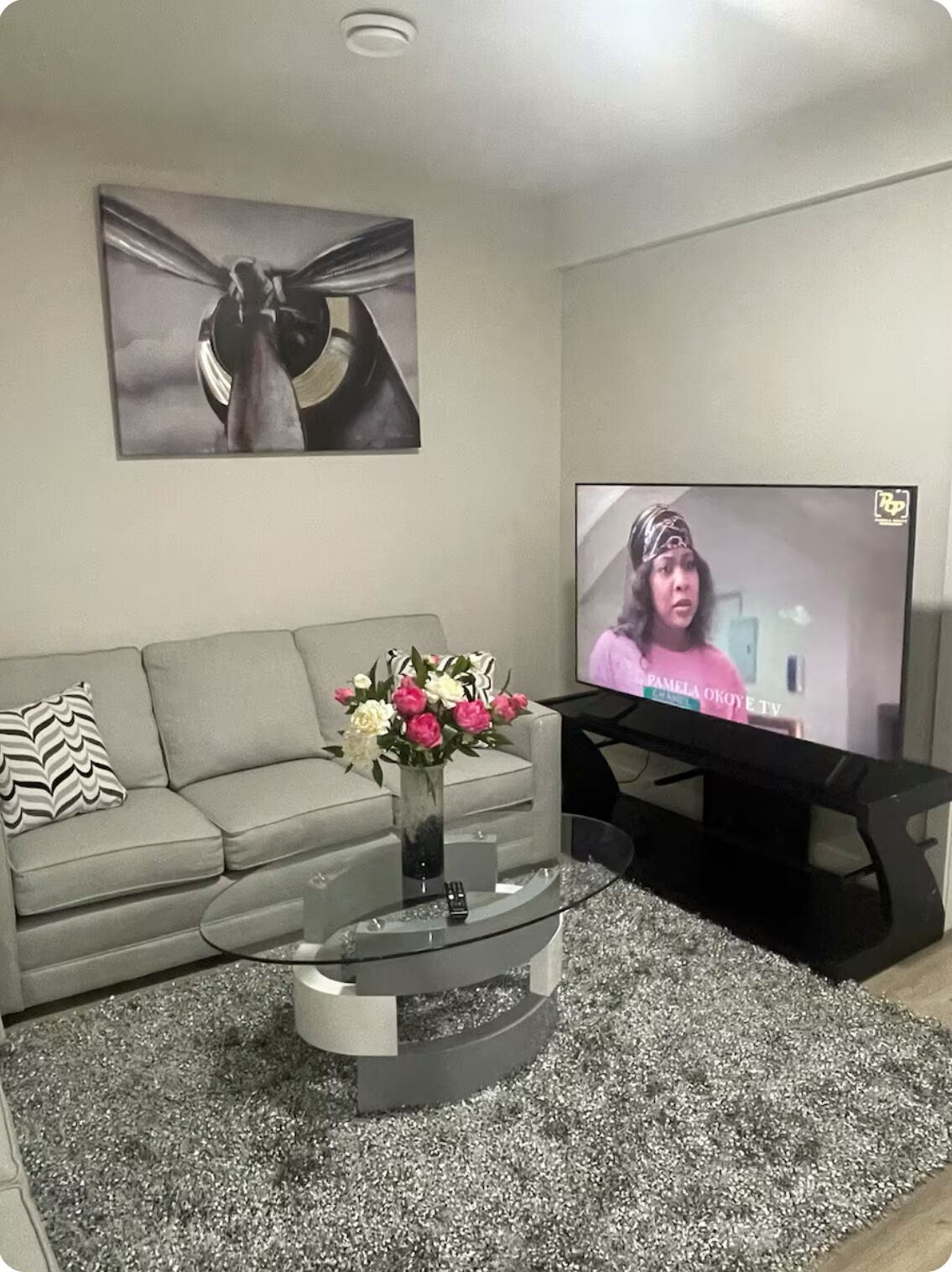 A living room with a grey sofa, a glass coffee table with a vase of flowers, a flat-screen TV, and a framed abstract painting on the wall.