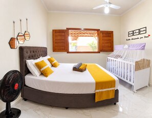 A bedroom with a bed, a crib, a standing fan, and a window with wooden shutters. - Mining House. Perfect for the relaxation your family deserves. (Nova Viçosa)