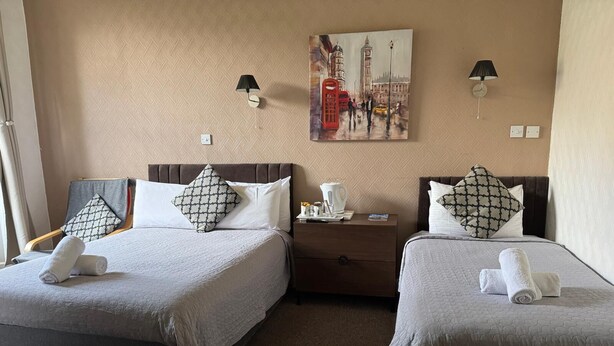 A hotel room with two beds, each with a pillow and a rolled towel. There is a nightstand with a lamp and a painting of a cityscape on the wall. - Sofia Rooms (Edinburgh)