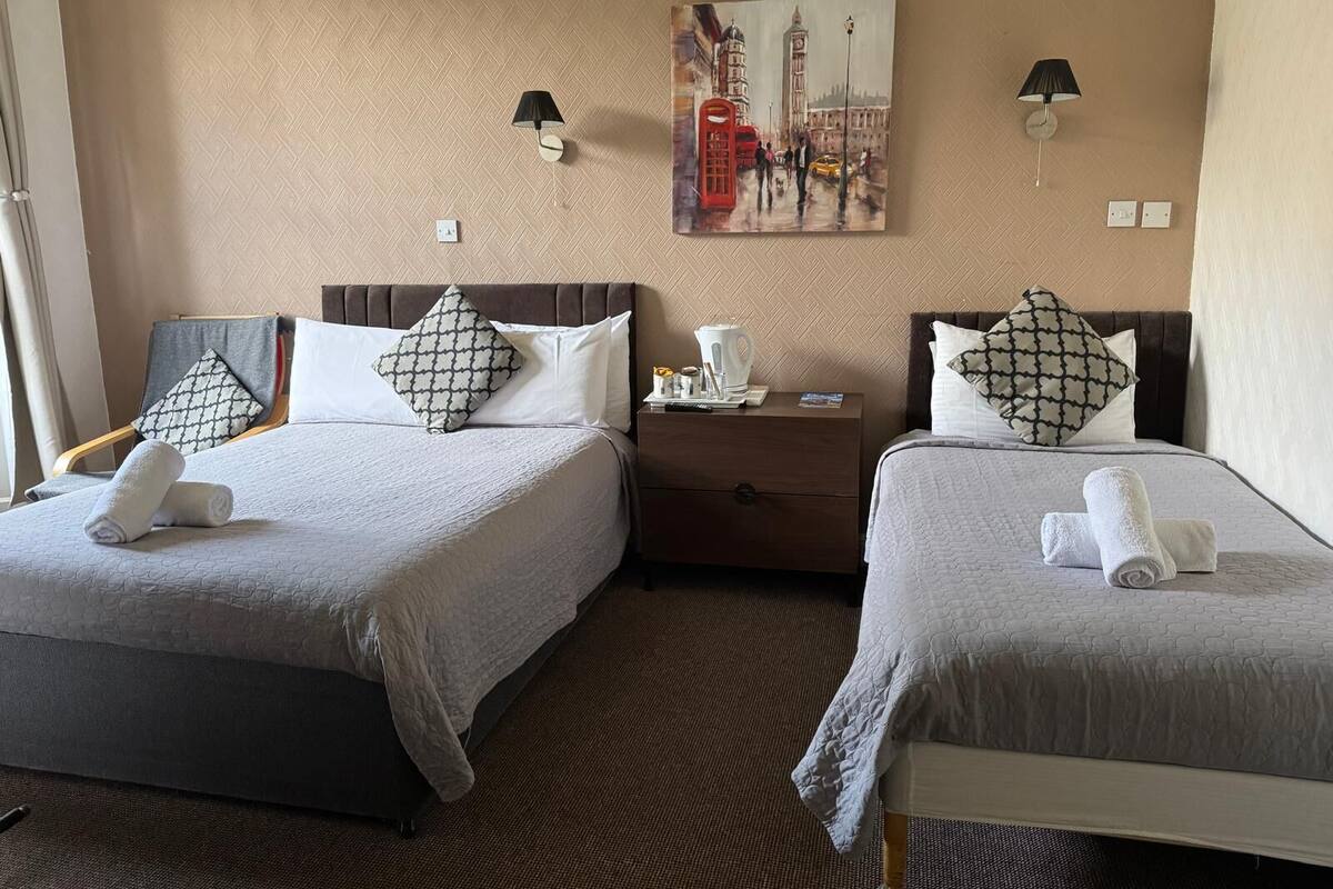 Habitación de hotel con dos camas, cada una con una almohada y una toalla enrollada. Se observa un mesita de noche con una lámpara y un cuadro de un paisaje urbano en la pared.