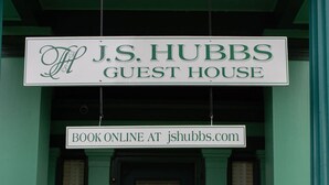 Un edificio verde con un letrero que dice "J.S. HUBBS GUEST HOUSE" y un letrero debajo que indica "RESERVE EN LÍNEA EN jshubbs.com". Hay dos sillas de mecedora de madera frente a la entrada.