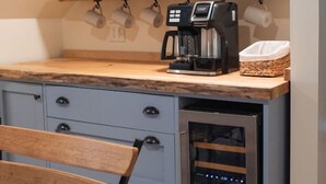 Una kitchenette compacta equipada con cafetera, horno empotrado y refrigerador pequeño. Se observan armarios de madera y una silla.