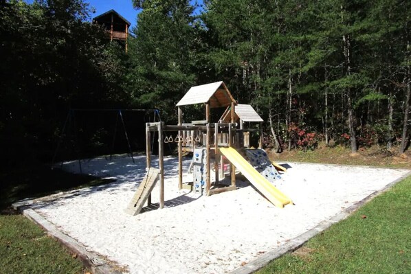 A wooden play structure with a slide and climbing elements, surrounded by a sandy area and trees. - Vista Ridge Retreat at Deer Ridge Mountain Resort (Gatlinburg)