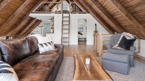 Een loft-achtige kamer met houten plafond, een leren bank, een grijze fauteuil en een houten salontafel.