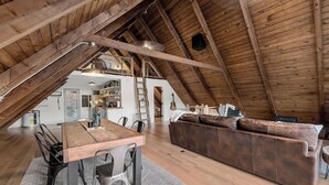Een kamer in zolderstijl met een houten plafond, een eettafel met stoelen, een leren bank en een keuken in de achtergrond.