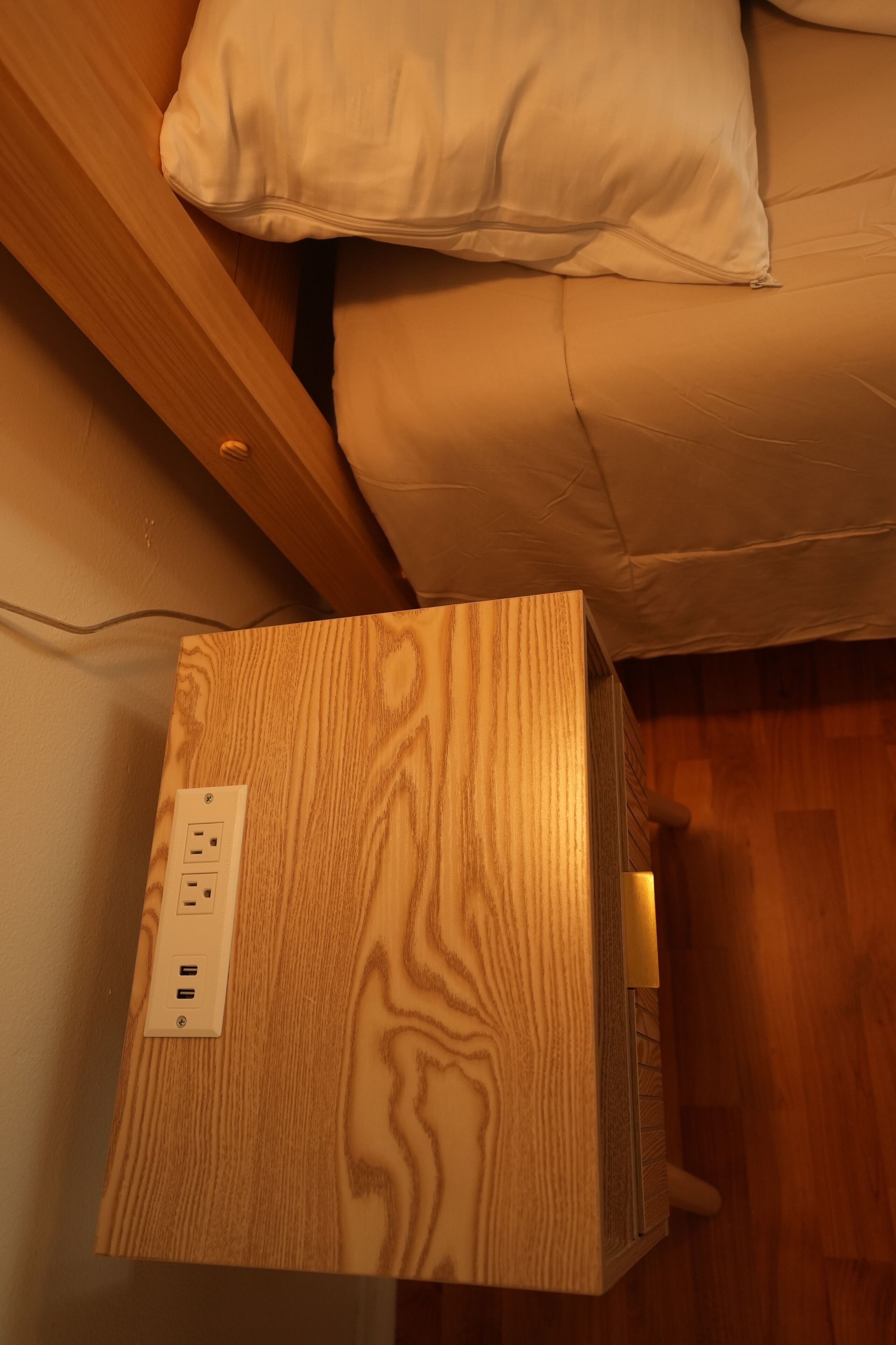 Una mesita de noche de madera con un enchufe, una lámpara y una cama con almohada blanca.