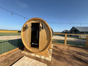 Eine holzartige, fassförmige Sauna mit einer Tür mit dem Schriftzug 'ALEKRO' und einem Metallzaun darum.