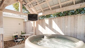 Ein Whirlpool unter einer Holzpergola, darüber ein Fernseher. Umgeben ist er von einem Holzzaun und einem kleinen Tisch mit Stühlen.
