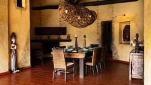 A rustic dining room with a wooden table set for four, surrounded by wicker chairs. A woven light fixture hangs from the ceiling, and a statue stands on the left.