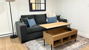 Una sala de estar moderna con un sofá gris oscuro, dos cojines azules, una mesa de centro de madera, una lámpara de pie y una alfombra estampada.