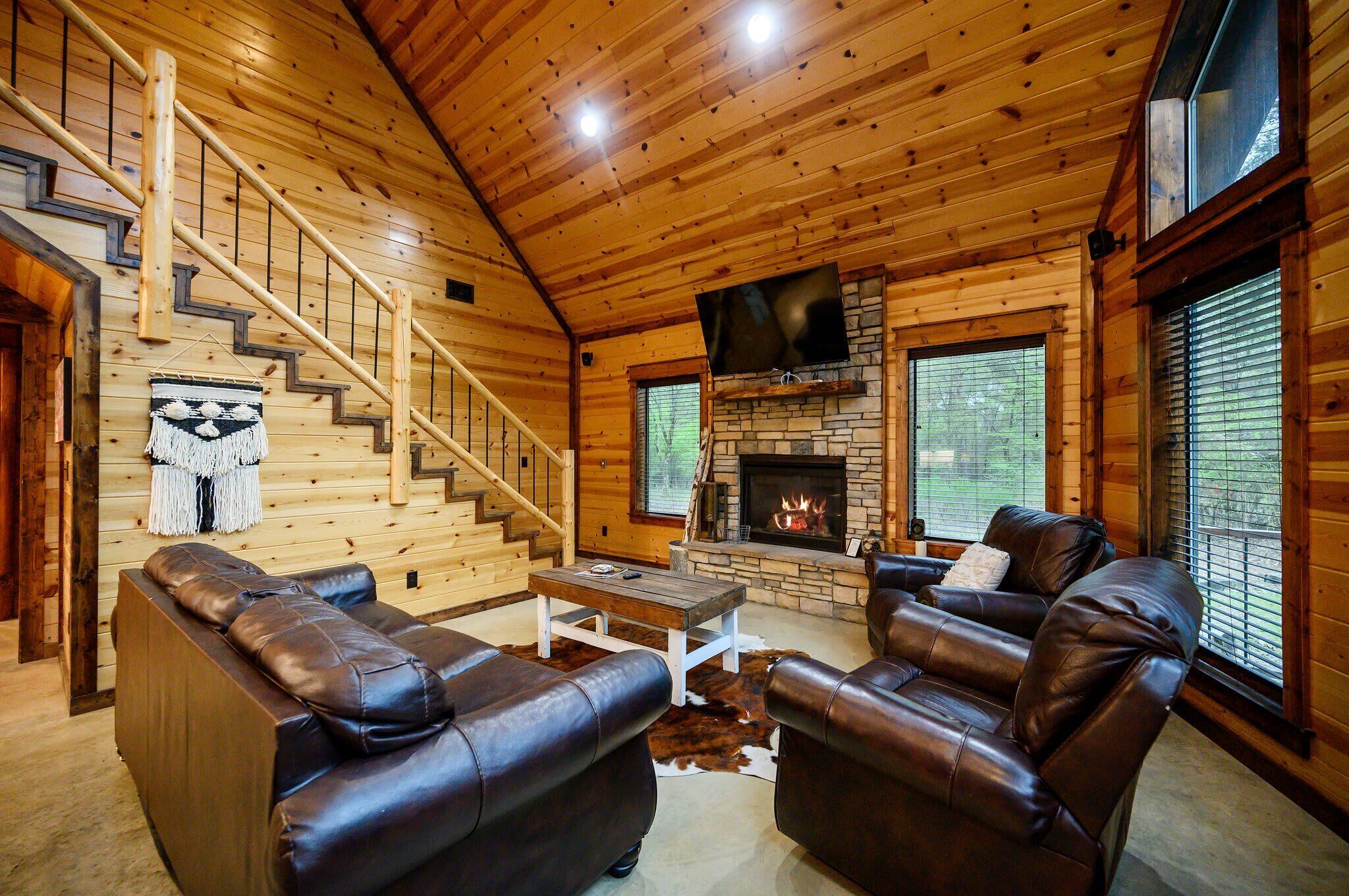 L’intérieur d’une cabane en rondins est agrémenté d’une cheminée, de canapés en cuir, d’un escalier en bois et d’un manteau de cheminée en pierre.
