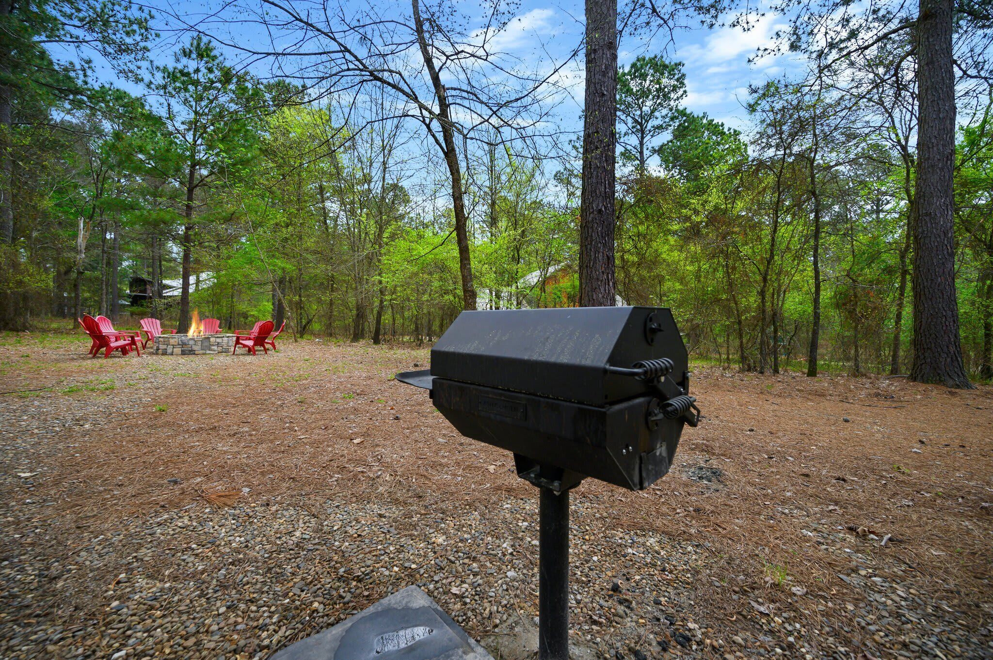 A barbecue grill in a wooded area with red chairs and a stone fire pit.