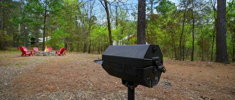 A barbecue grill in a wooded area with red chairs and a stone fire pit.