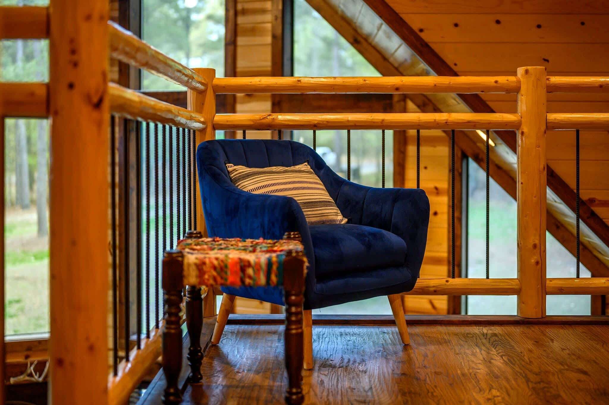 Una stanza con struttura in legno, una poltrona blu e un tavolino, una finestra con vista sul verde, e un soffitto spiovente.