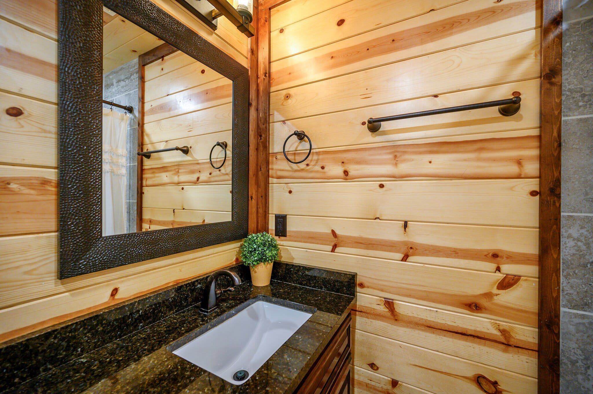 A wooden cabin bathroom with a granite countertop, a white sink, a mirror, a towel rack, and a small potted plant.