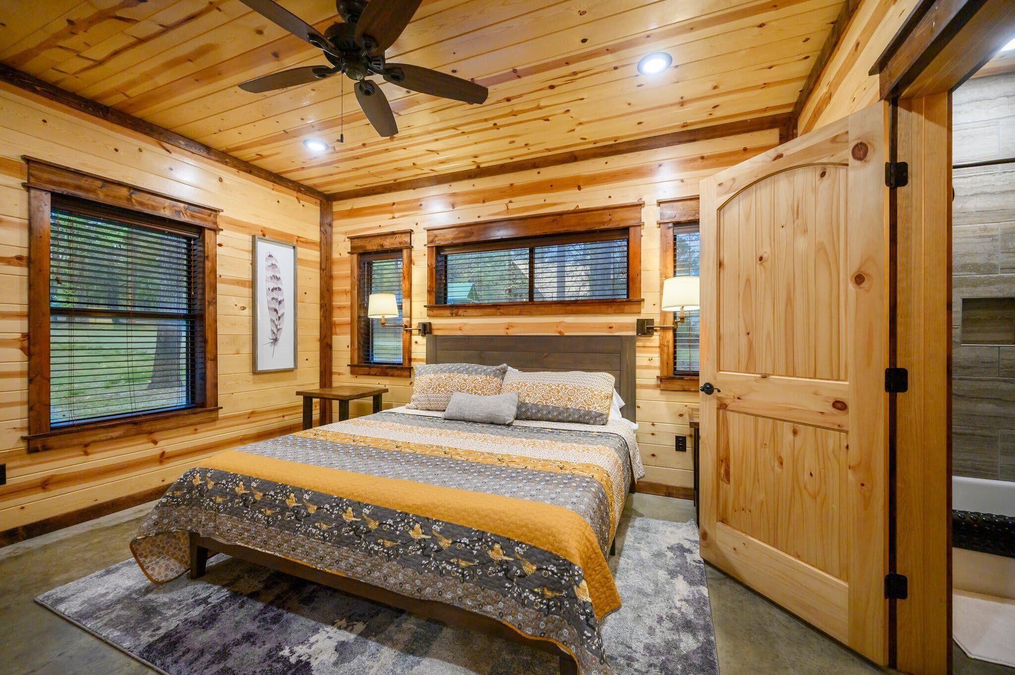 A wooden cabin bedroom with a bed, two bedside tables, a ceiling fan, and a large wooden door.