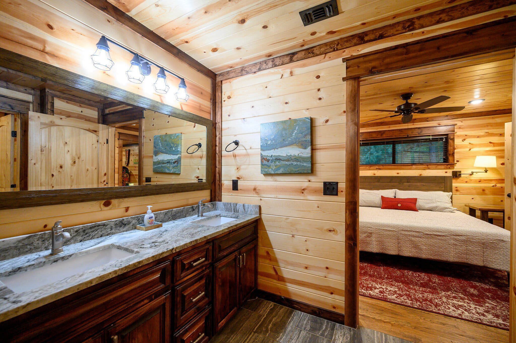 L’intérieur d’une cabane en bois comprend une salle de bain équipée d’un lavabo, d’un meuble-lavabo et d’un miroir. On y trouve un lit avec un oreiller rouge et un ventilateur de plafond au-dessus.