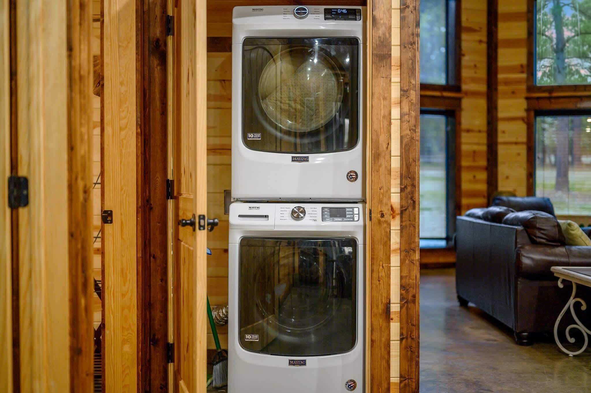 A stacked washer and dryer unit in a wooden cabin with a couch and a window in the background.