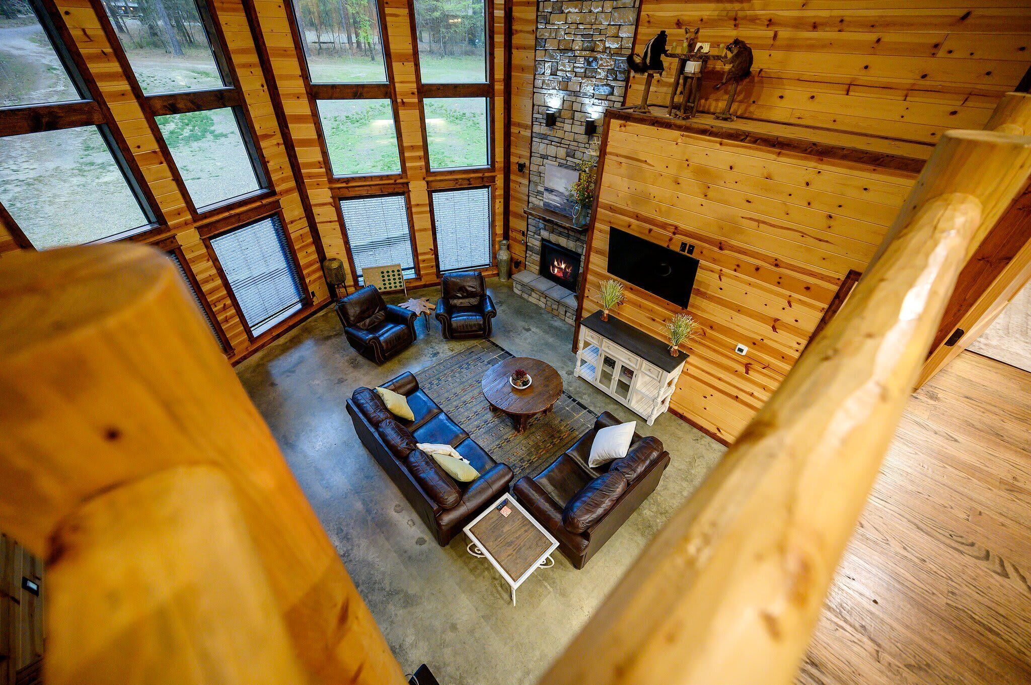 A log cabin interior with a stone fireplace, large windows, and a cozy seating area.