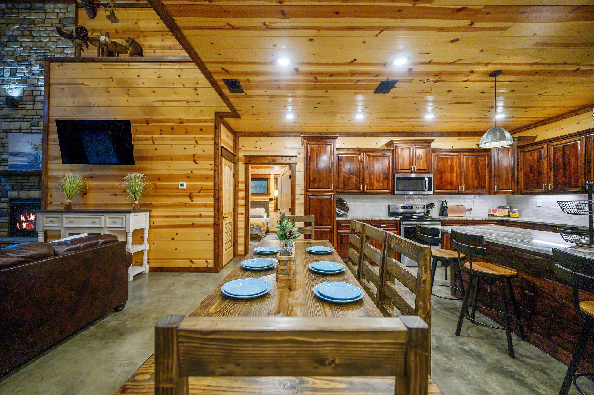 A wooden interior with a dining table set for six, a kitchen area with wooden cabinets, a stone wall, and a fireplace.