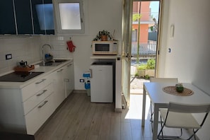 A compact kitchen with white cabinets, a microwave, a small refrigerator, and a dining area with a table and chairs. - CN009 - Cesenatico, new two-room apartment with garden and parking space (Cesenatico)