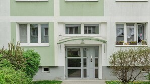 Un immeuble à plusieurs étages, doté d’une façade verte et blanche, d’une entrée centrale avec une porte et une plaque de numéro, et de nombreuses fenêtres ornées de rideaux.