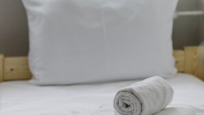 Un lit bien fait, avec une serviette blanche roulée et un oreiller blanc, dans une pièce dotée d’une fenêtre et d’un rideau.