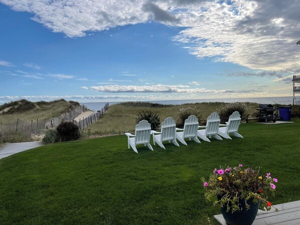 Zona verde con sillas Adirondack blancas dispuestas en fila, con vista a una playa y dunas.