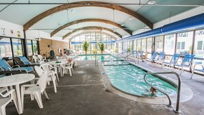 Une piscine intérieure dotée de chaises longues et de tables, entourée de grandes fenêtres et de poutres en bois.