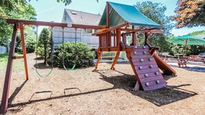 Une aire de jeux en bois avec un toboggan et des balançoires, entourée d’arbres et avec une maison en arrière-plan.
