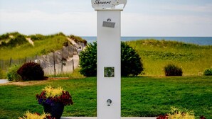 A white structure with a sign reading "Shower" and a decorative element resembling a horse's head. It is placed on a wooden platform with two potted plants with red and yellow flowers. - Oceanfront 1-Bedroom Condo w/ Balcony Views (Dennis Port)